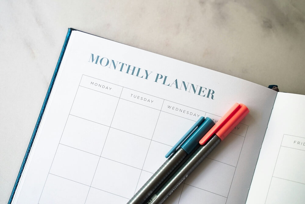 An open page of a monthly planner book, with a blue pen and orange pen resting on the open page. 
