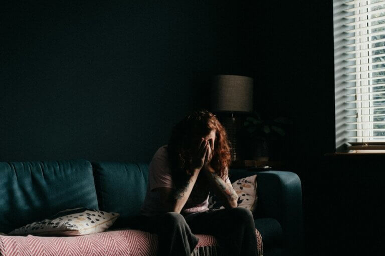 A woman with wavy red hair and tattooed arms sits on a couch in a dark living room, dimly lit by a nearby window. She has her hands up to her face, rests her elbows on her knees, and looks distraught, though we can't see her face.