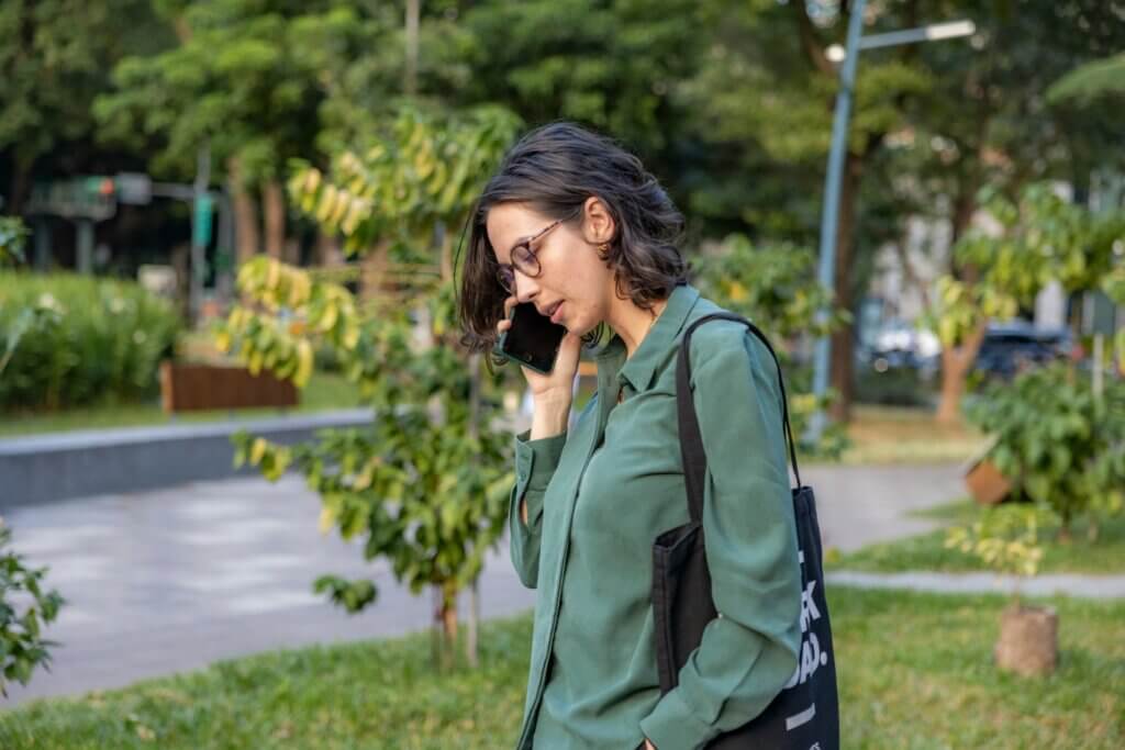 A woman with a wavy brown bob haircut, brown glasses, and a microsuede green collared shirt is on a phone call in a park. She holds a cell phone to her ear and appears to be speaking.