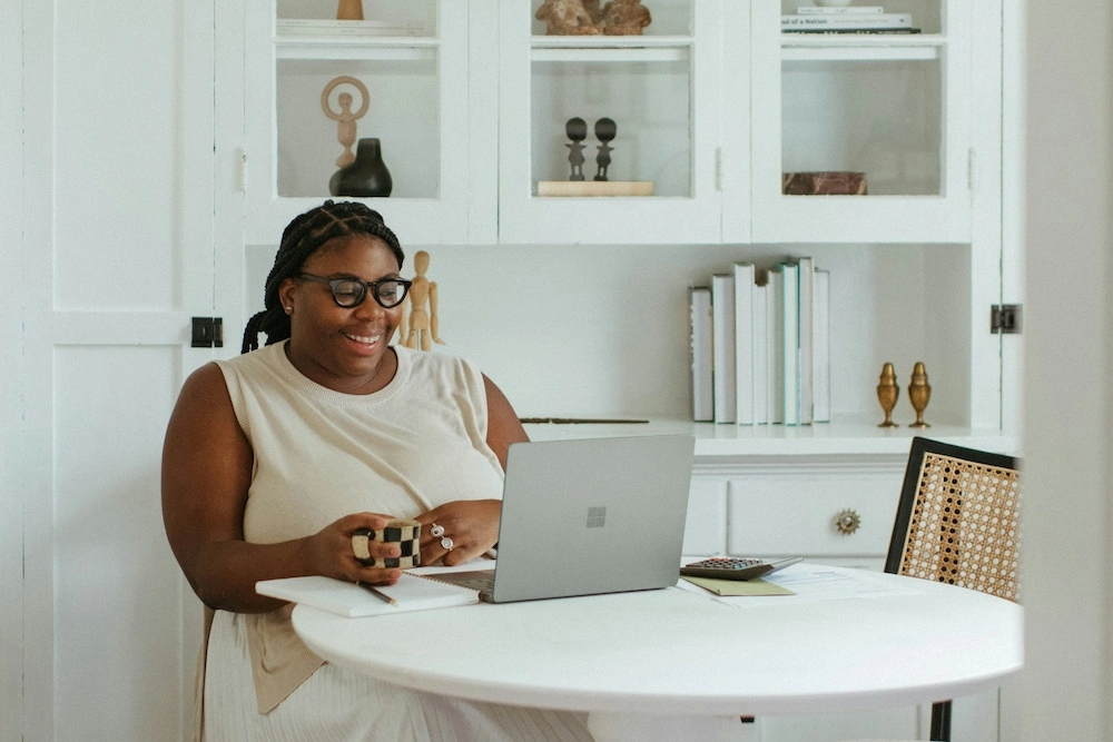 A Black woman in a cream-colored sleeveless shirt and white linen pants sits at a white circular coffee table in a white dining room setting. She wears black cateye glasses and has long hair in braids tied back. She's laughing, looking at the screen of a silver Surface laptop as though on a video conference with someone. She holds a smal ceramic black-and-white checkered coffee mug, with large decorative white rings on her other hand. 