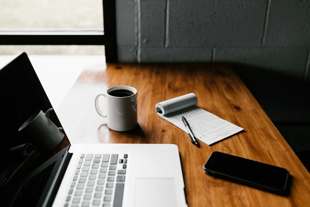A window seat in an office with painted gray brick walls. On a slim wooden tabletop sits a MacBook laptop, a white coffee mug filled with black coffee, a notebook open to a filled page with a black pen resting on it, and a black phone. 