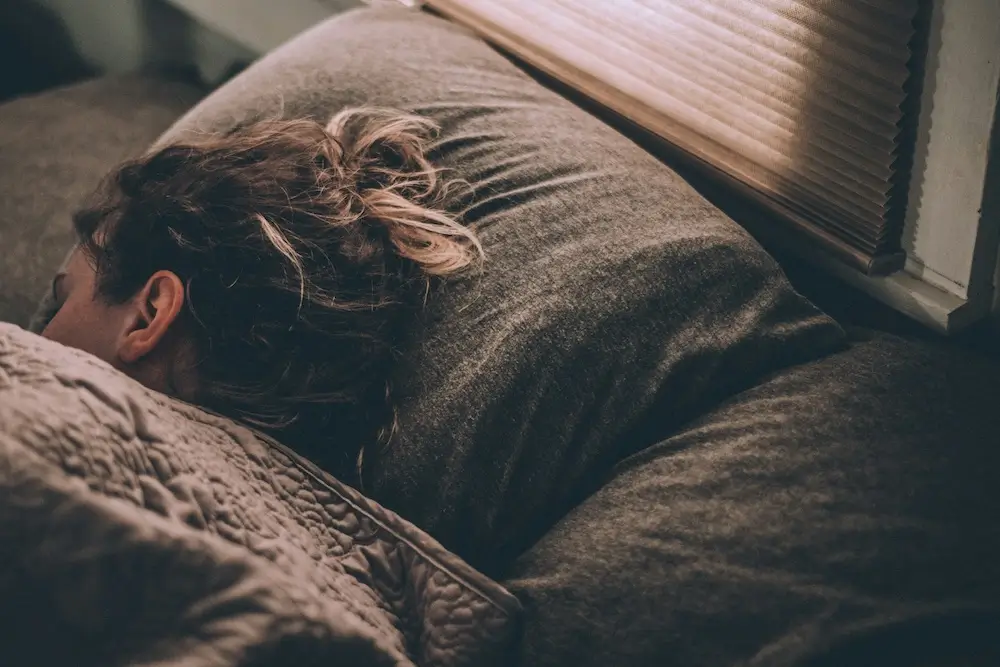 A morning scene of a white person with brown hair and dyed blonde tips nestled into a gray jersey pillow, under a gray quilt. Their bed is against a window with white trim and a gray accordion window shade. They seem to be sleeping.