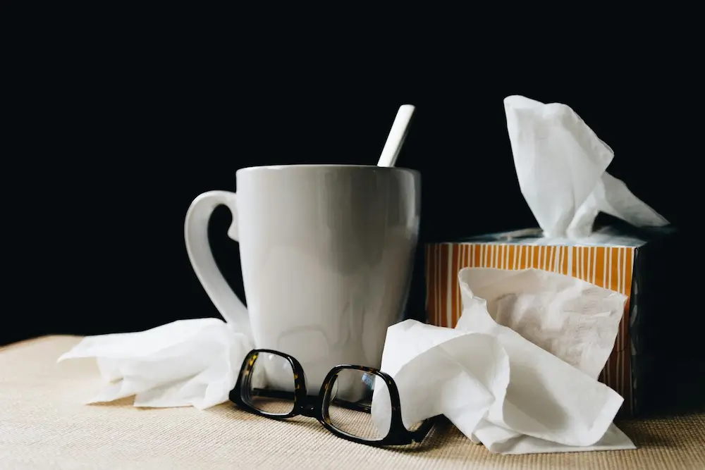 White coffee mug with stir stick, black tortoiseshell glasses turned upside down, and a box of tissues sit on a woven fabric table top in front of a black background. A few loose tissues surround the coffee mug. All together, it's a scene of someone who's home sick.