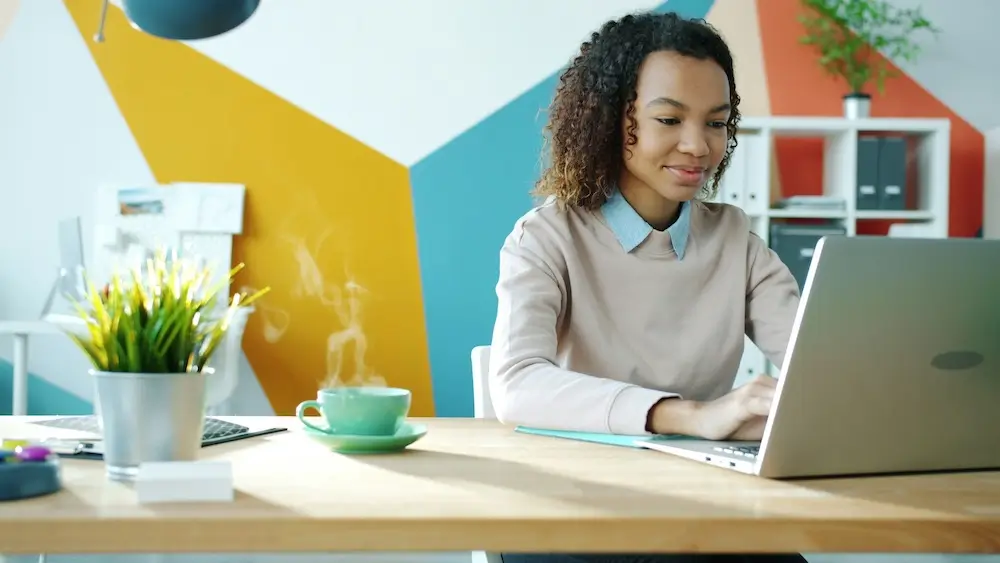 Young Black woman sits at a light wood desk in a bright office with a colorful geometric mural on the wall behind her. She smiles as she types on a laptop; a steaming teal cup on a saucer sits on the desk beside her.