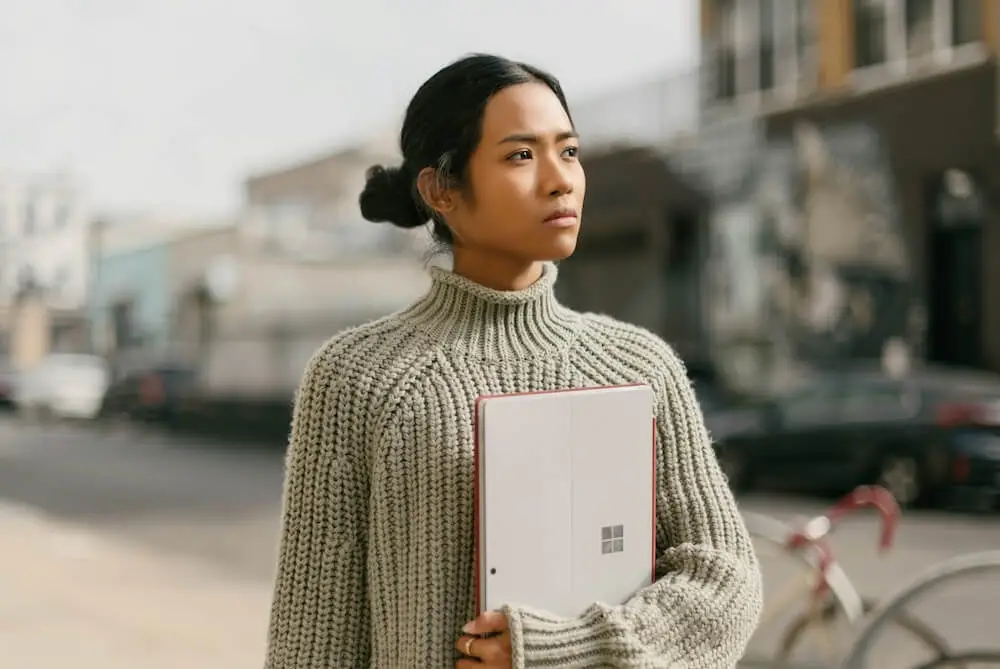 A young Southeast Asian woman stands on a city street, wearing a chunky off-white cable-knit sweater and holding a Microsoft tablet with one hand, pinned against her torso. She looks off into the distance as though thinking, but appears confident.