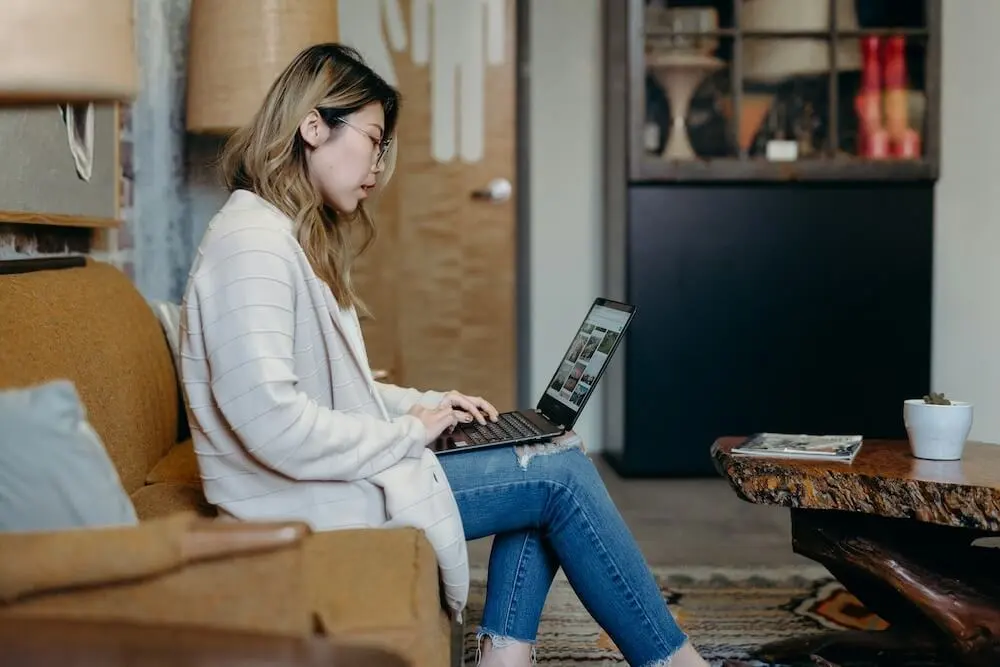 An Asian woman with bleached blond hair sits in a living room on a caramel-colored couch. She wears a long beige cardigan and skinny jeans. She's working on a laptop that rests in her lap.
