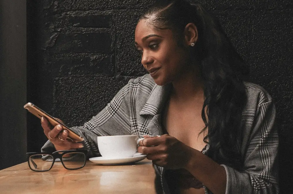 Black woman with long wavy ponytail wears a black and white patterned blazer with sleeves rolled up. She sits in a coffee shop with black brick walls, one hand on the handle of a white coffee mug and the other holding her phone. She leans on a live-edge wood countertop and looks at her phone, smiling slightly. 