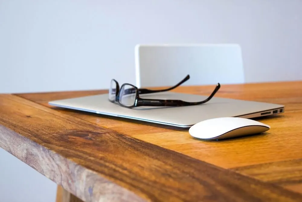 A wood tabletop with a closed MacBook; a pair of black rectangular glasses rest upside down on top of the laptop. A white wireless mouse sits next to the laptop.