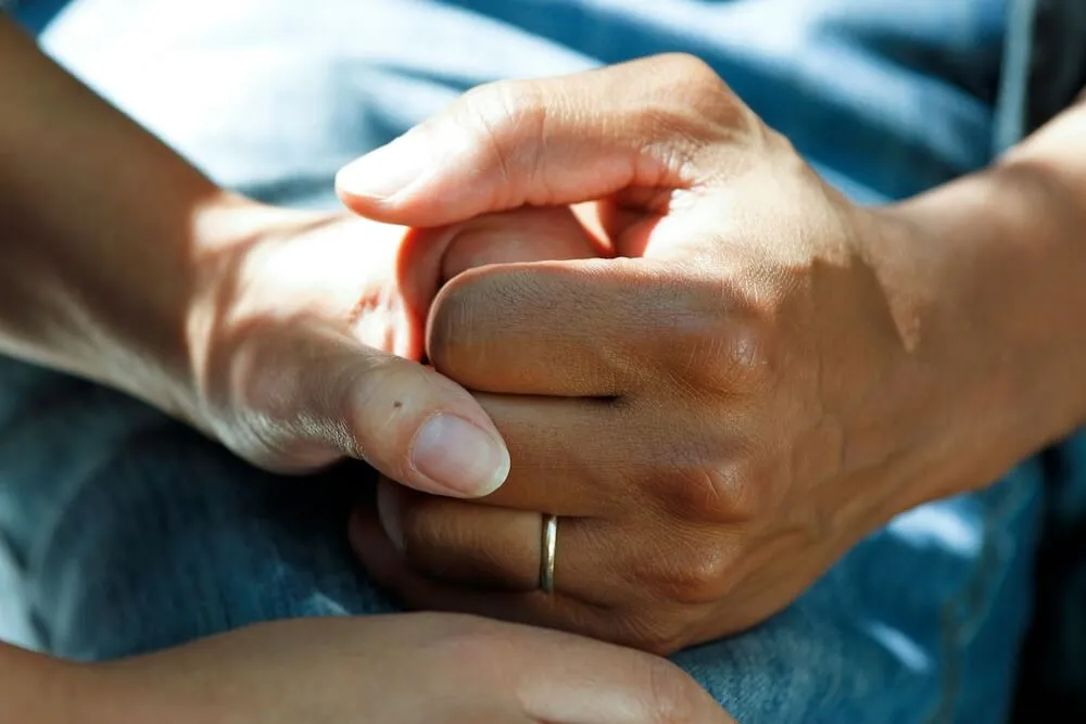 A close-up of two people's hands, with lightly tan skin. One person is holding the other's hand. The hand grasping on the outside has a thin white-gold ring; the hand being grasped holds back with their thumb, and there's a small spot just below their thumbnail. The blue denim background appears to be one person's jeans.
