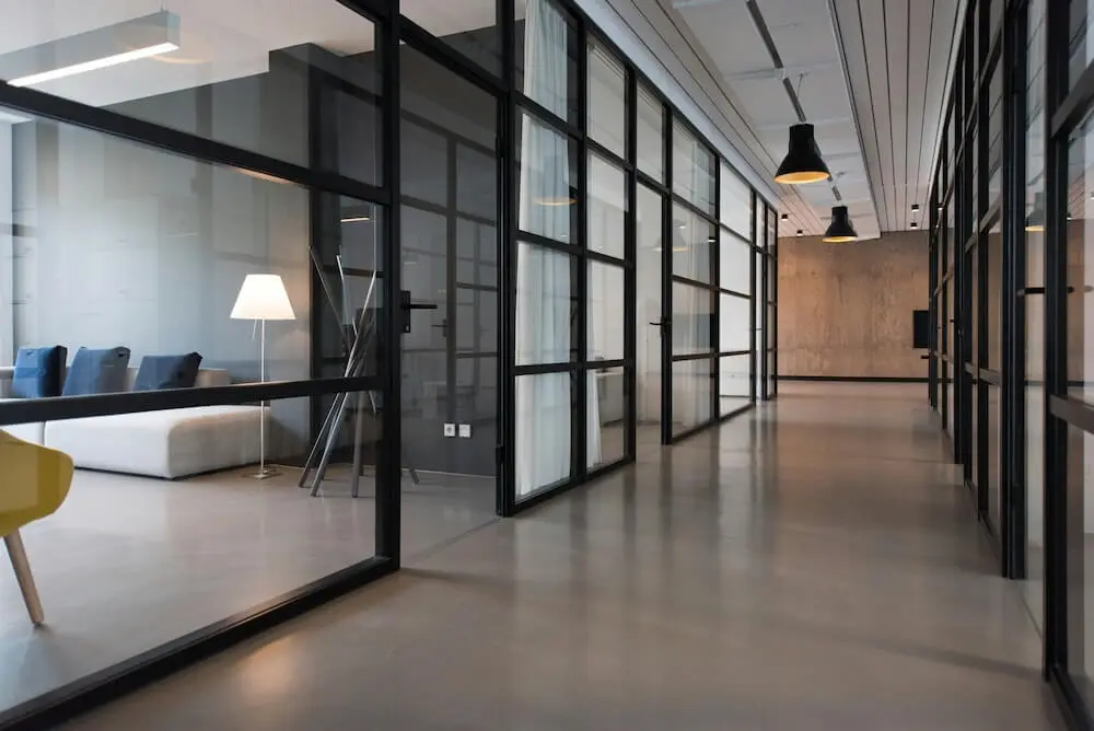 A hallway in a modern corporate office with floor-to-ceiling window walls, lined with black frames. A muted brown wooden floor spans the whole scene, and some modern office furniture (a chair, white couch, and large lamp) are visible in one room.