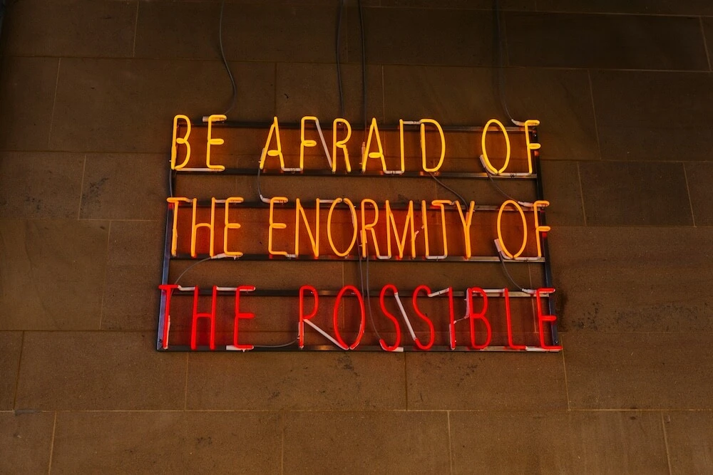 A neon sign mounted on a concrete wall reads, in all capital letters, BE AFRAID OF THE ENORMITY OF THE POSSIBLE, with "BE AFRAID OF" on the first line in yellow, "THE ENORMITY OF" on the second line in orange, and "THE POSSIBLE" on the third line in red.