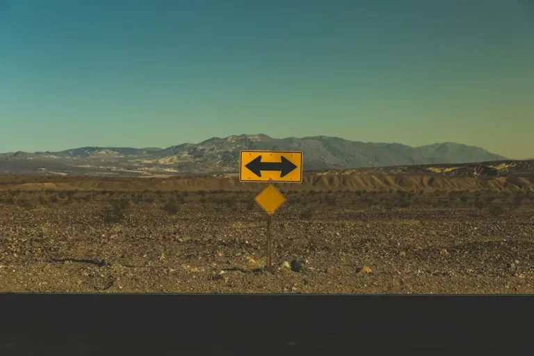 A yellow road sign with a black two-way arrow at the edge of a black asphalt road in the desert, indicating drivers can turn left or right but can't go straight