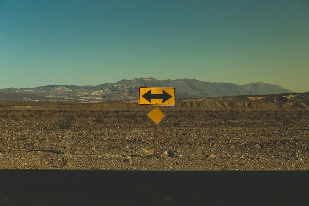A yellow road sign with a black two-way arrow at the edge of a black asphalt road in the desert, indicating drivers can turn left or right but can't go straight