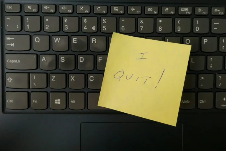yellow sticky note reads "I QUIT!" in blue ballpoint ink. it's stuck to a black computer keyboard