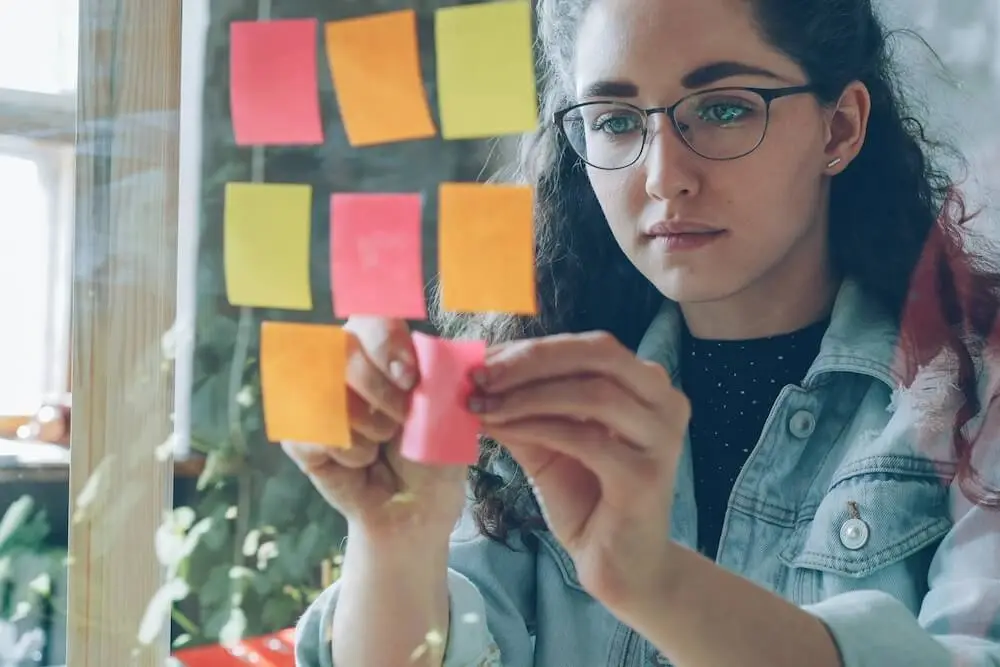 A white woman in her 20s with curly brown hair past her shoulders wears a black t-shirt with light chamay shirt layered over it. She looks very focused, sticking multi-colored sticky notes onto a glass wall as though in a planning session.