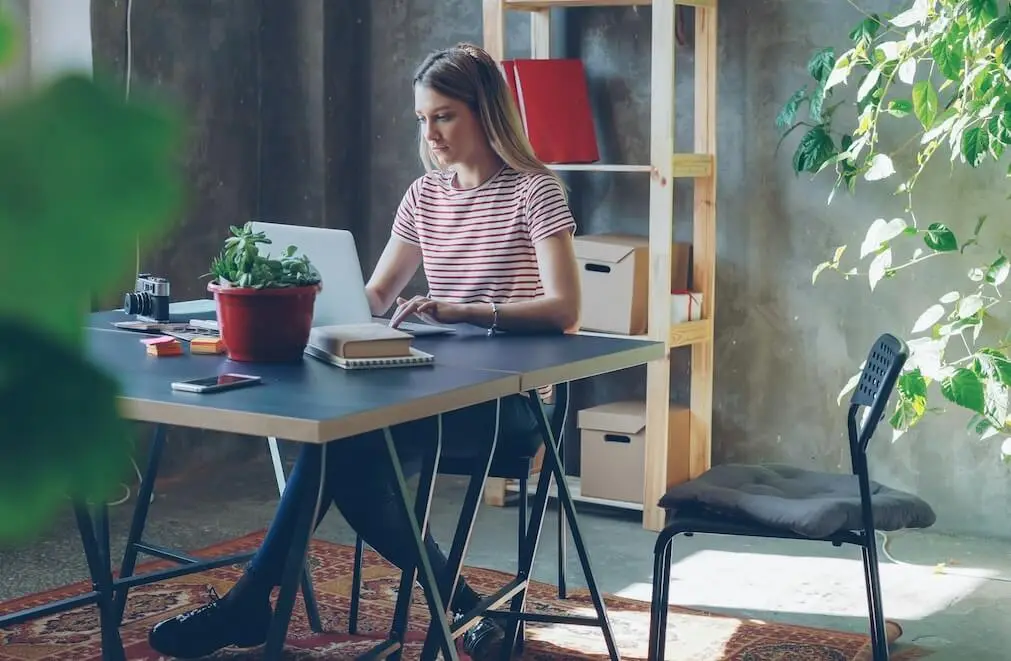 A young white woman with long, straight, blond hair sits at a desk in a modern, minimalist, open office with concrete walls and floors. She's working on a laptop in a sunlit area. There's a plant in a red planter on the desk near her, and a plain pine shelving unit behind her.