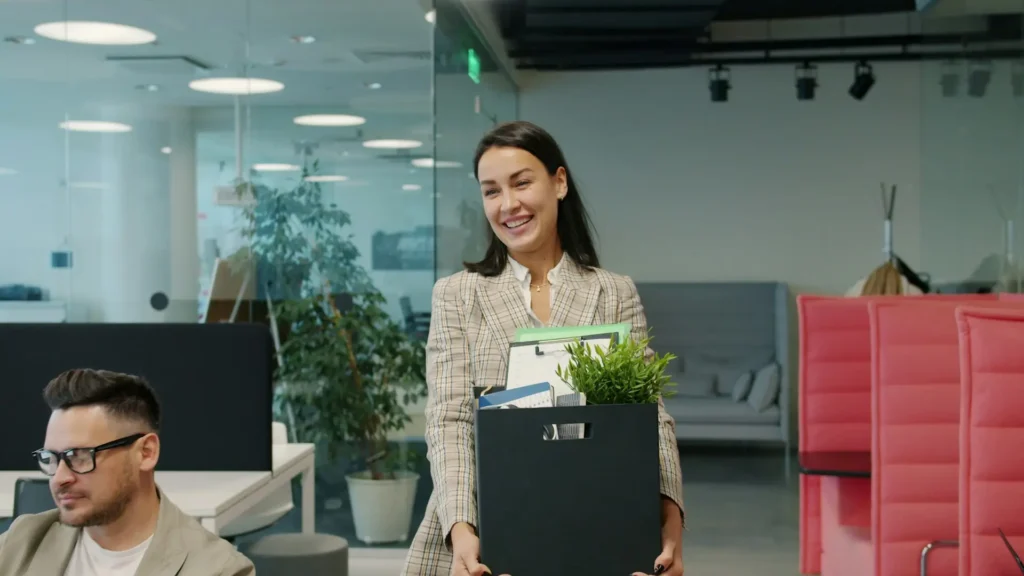 white woman with straight dark brown hair carries a black box filled with a plant and office supplies. she's walking through a modern office, so it looks like she's leaving her job. she smiles and looks at someone out of the frame.