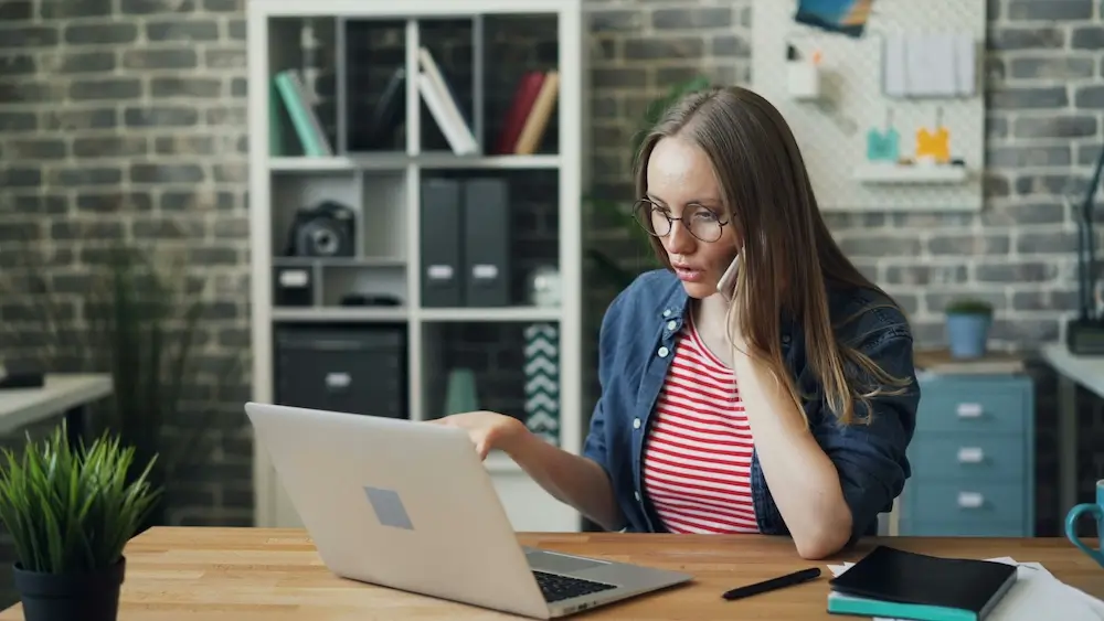 A white woman in her 20s with long blonde-brown hair, round black wire glasses, a pink-and-red striped shirt, and an unbuttoned layered chamay shirt sits at a wooden desktop in an office setting. She's speaking on the phone and looking at her laptop with one hand upturned and gesturing toward the screen, as though seeking an answer to a question about something she's looking at.