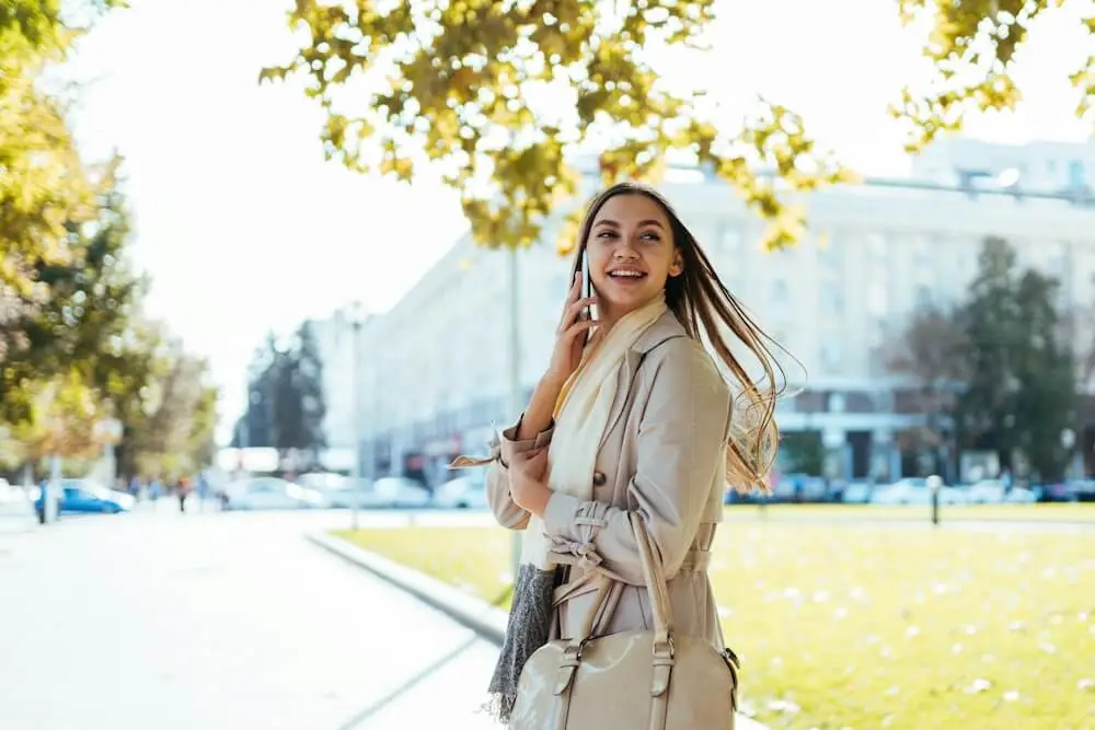 A young white woman with long brown hair looks over her left shoulder as she walks down a city street, near a grassy lawn and approaching a large building. She wears a khaki blazer and white scarf, holding a khaki-colored purse on her arm. She's talking on a cell phone and smiling. It's a sunny day, and a leafy tree branch illuminated by the sun hangs above her.