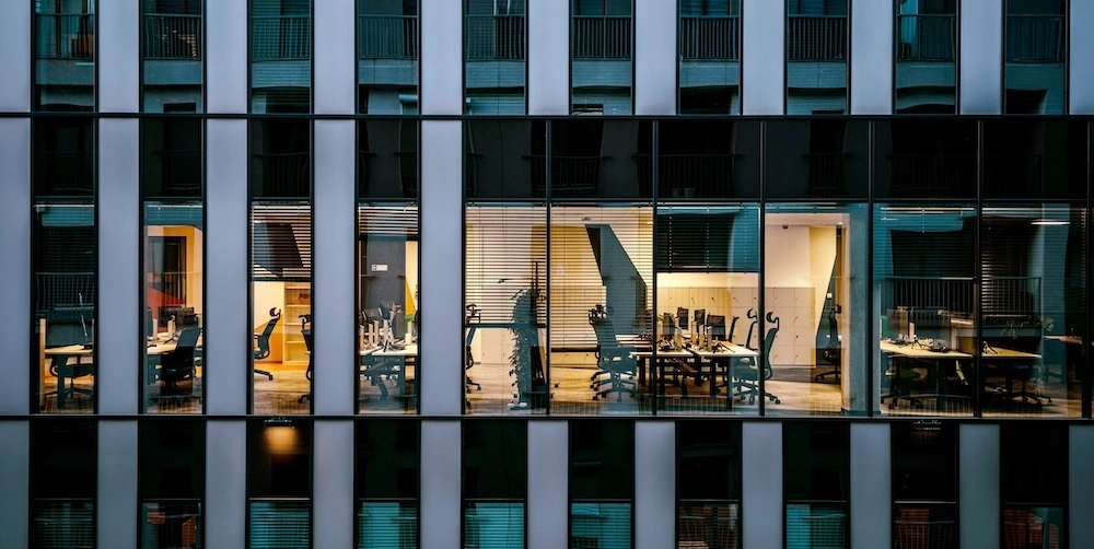 external view of an office building, showing several conference rooms and offices through the windows. the overall mood is bleak; shades are drawn to varying heights and most of the lights on the floors above and below are turned off. overall, it's a bleak corporate office