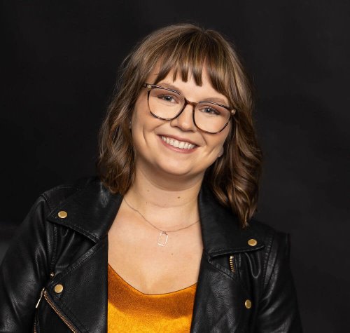 Coach Lucy Todd smiling with head tilted, wearing a black motorcycle jacket with rose gold accents, a metallic orange shirt, a silver pendant necklace, and brown glasses